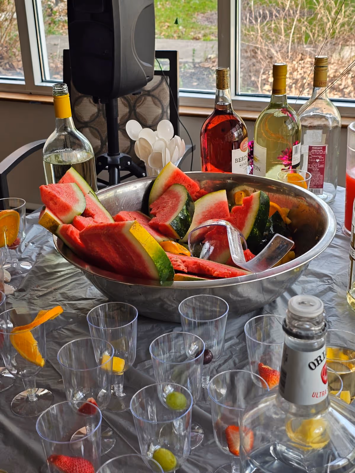 A table covered with a silver tablecloth displaying a large metal bowl filled with watermelon slices and some orange slices. Surrounding the bowl are multiple clear plastic cups, each containing a piece of fruit like grapes or strawberries. Behind the bowl are several bottles of wine and other beverages, along with a container holding white plastic spoons. The setting appears to be indoors near a window with a view of an outdoor area.