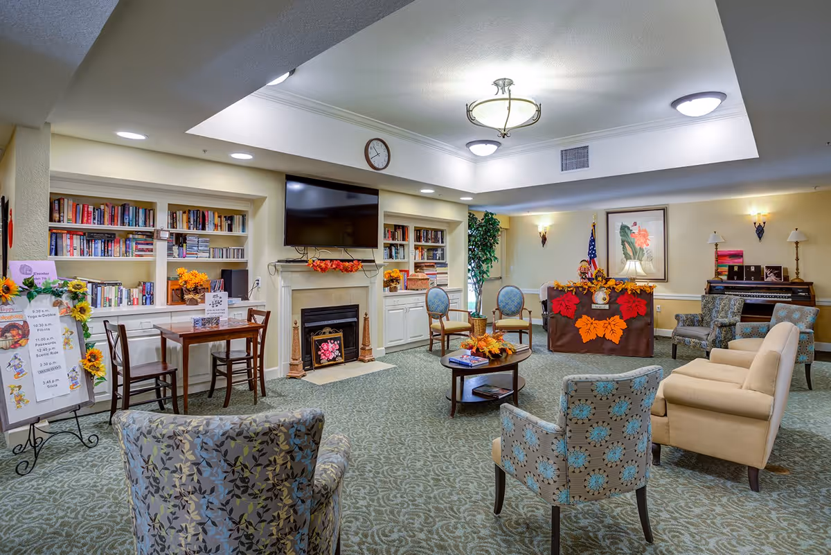 A cozy senior living facility common area with a fireplace, a wall-mounted TV, bookshelves filled with books, and various chairs and sofas arranged around a coffee table decorated with autumn leaves. There is a small table with chairs near the bookshelves, a decorated bulletin board with event information, and a piano in the corner. The room is warmly lit with ceiling lights and wall sconces.