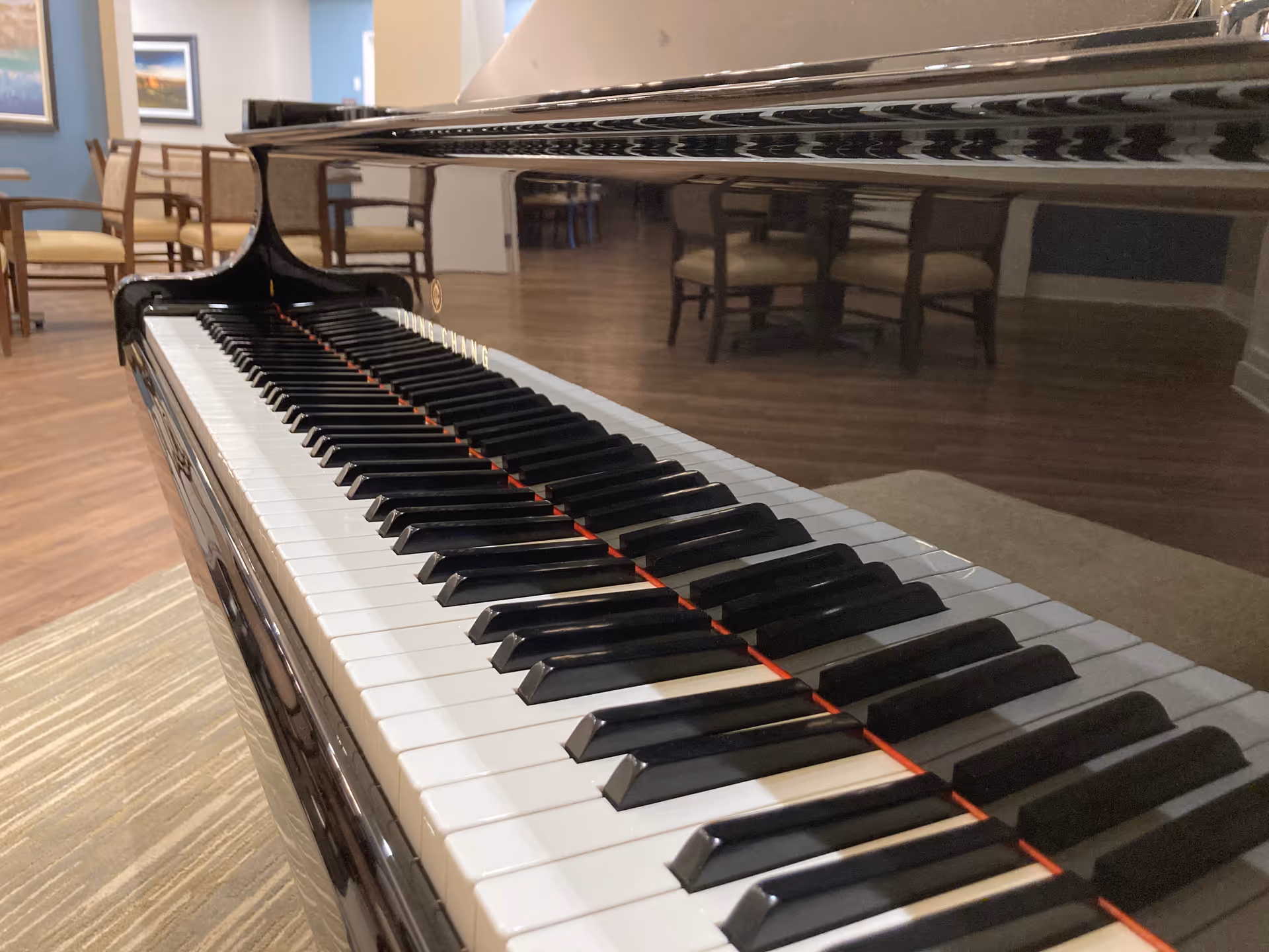 Close-up view of a black grand piano keyboard in a room with wooden flooring and several chairs and tables in the background.