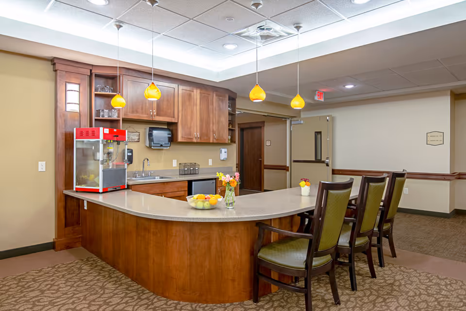 A communal kitchenette with a curved countertop, high chairs, wooden cabinets, pendant lights, and a popcorn machine.