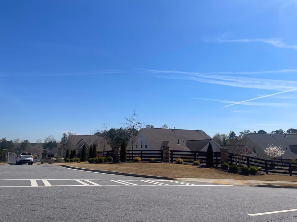 Street view of a gated residential community with houses, a wooden fence, and a car under a clear blue sky.