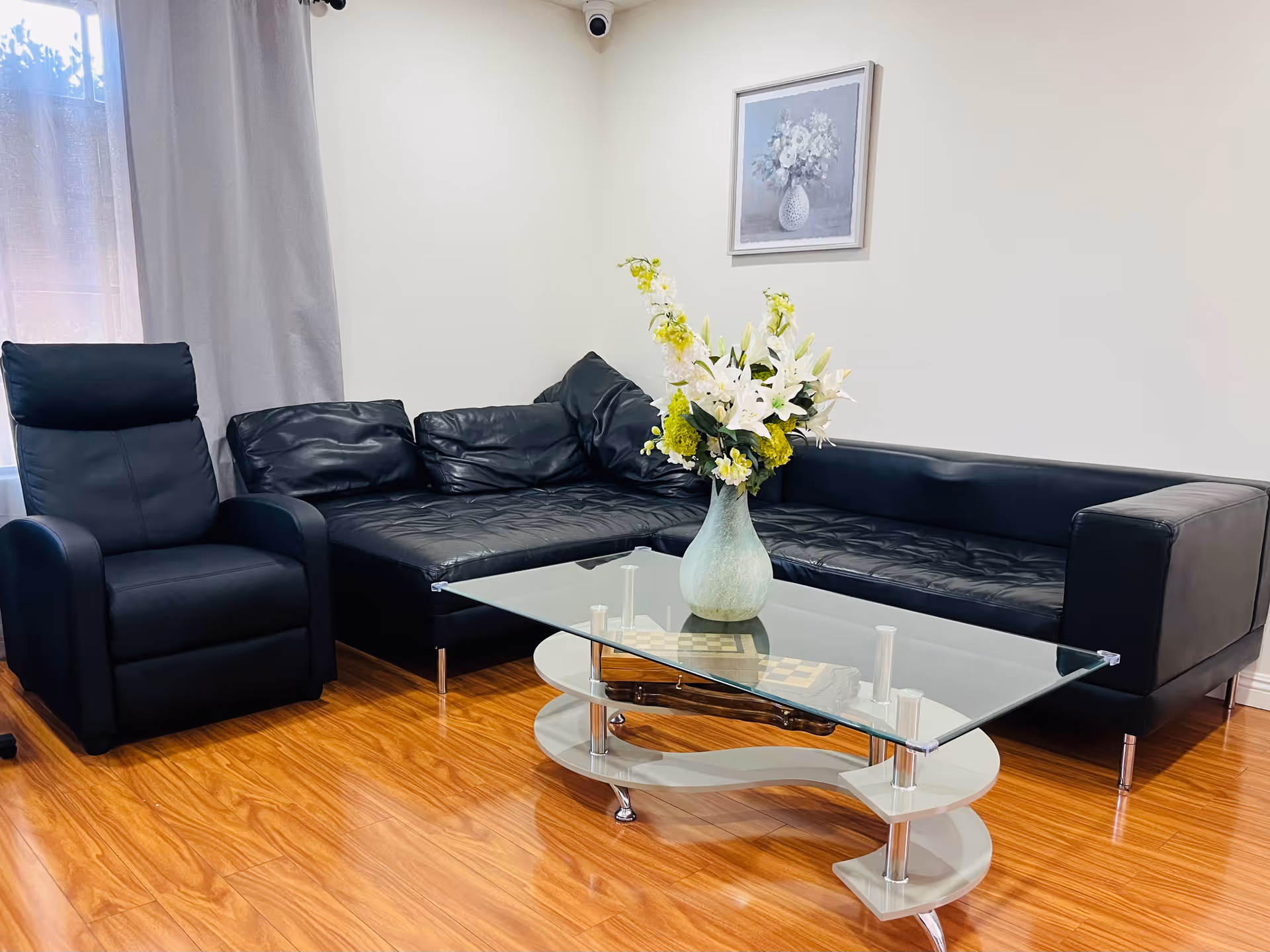 A modern living room with a black leather sectional sofa and a matching black armchair. A glass coffee table with a white base holds a vase of white and yellow flowers. The room has wooden flooring, a window with light curtains, and a framed floral painting on the wall.