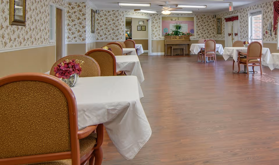 A dining room with several tables covered with white tablecloths, each accompanied by two chairs. Small floral centerpieces are placed on the tables. The room has floral wallpaper, wooden flooring, and a piano against the far wall with plants and decorations on top. Windows on the right side allow natural light to enter.