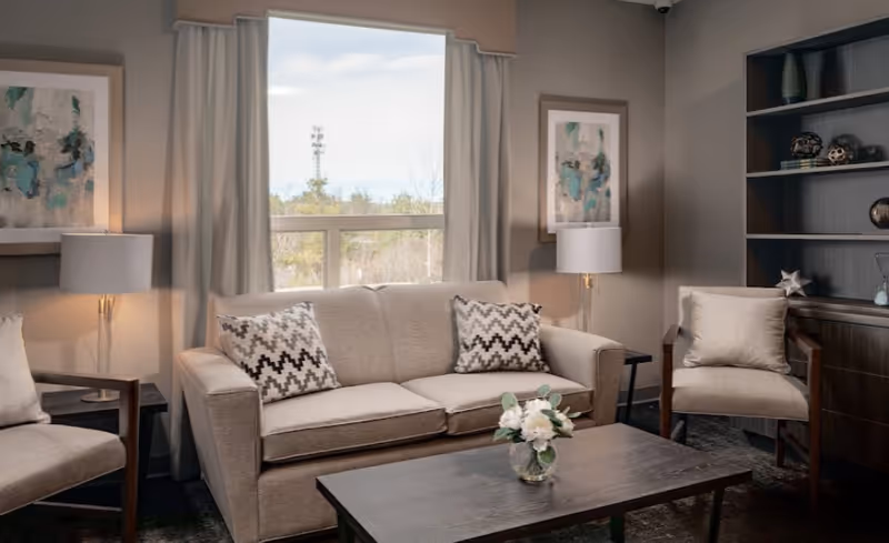 A cozy living room with a beige sofa and two matching armchairs, each with a cushion. There is a dark wooden coffee table in the center with a small vase of white flowers. Two table lamps with white shades are placed on side tables next to the sofa. The room has a large window with beige curtains, and two abstract paintings hang on the walls. A built-in bookshelf with decorative items is visible on the right side.