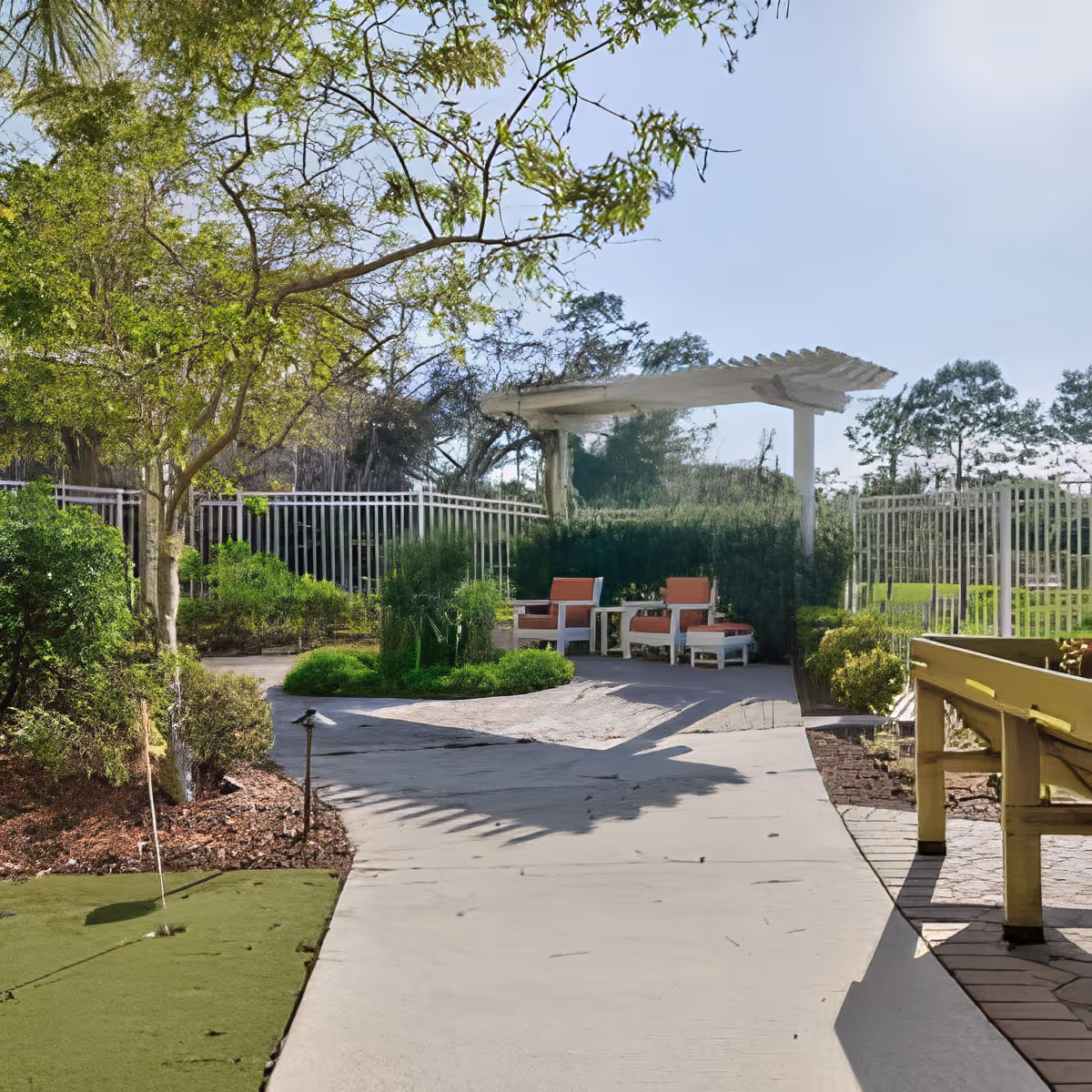 Outdoor garden area with a paved walkway, green bushes, trees, and a white pergola with two orange cushioned chairs and a small table underneath. There is a putting green on the left side and a wooden bench on the right side.