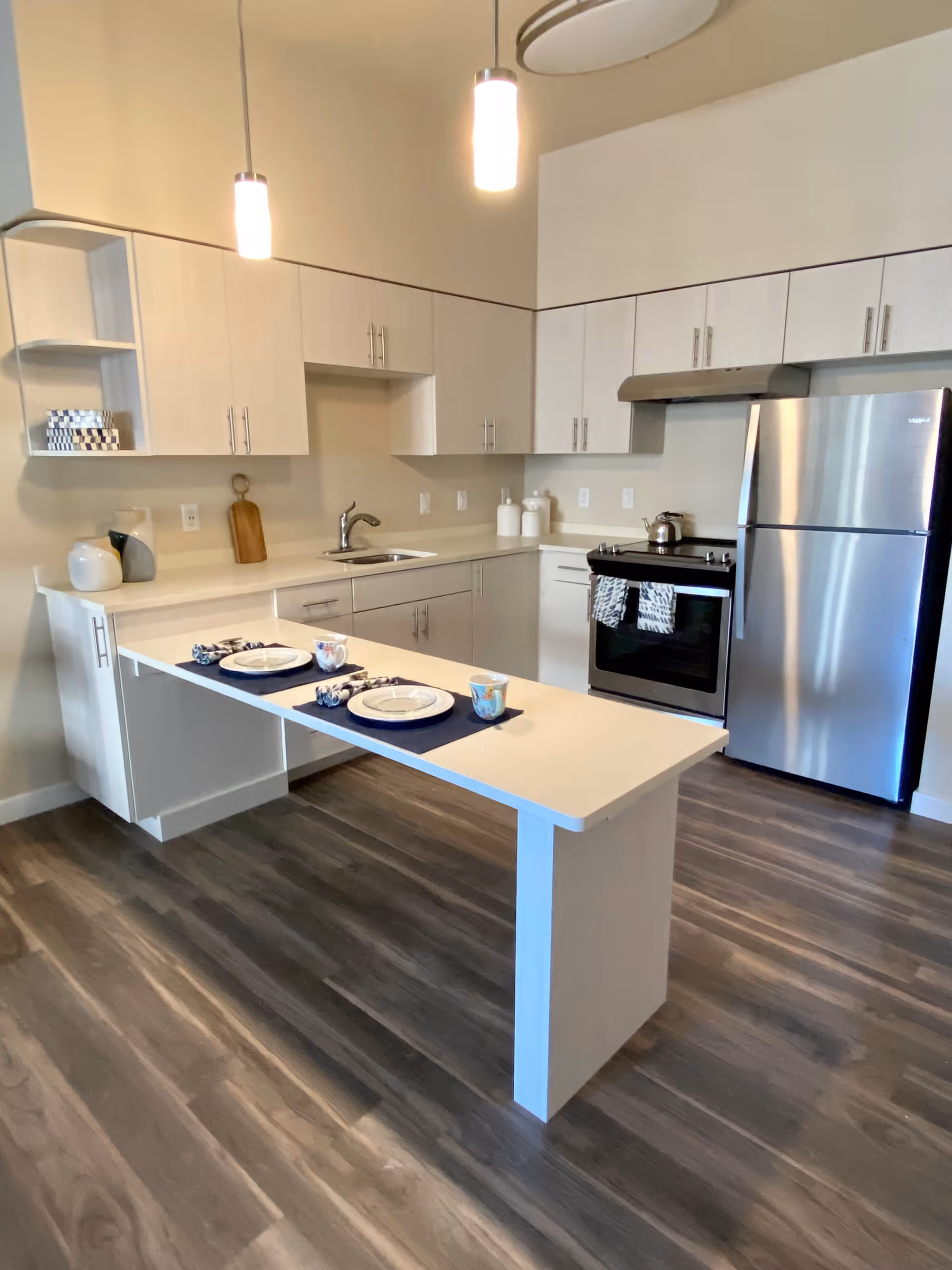 Modern kitchen with light wood cabinets, stainless steel refrigerator and oven, a sink, and a white countertop island set with two place settings including plates, cups, and napkins. The floor is dark wood, and two pendant lights hang from the ceiling.