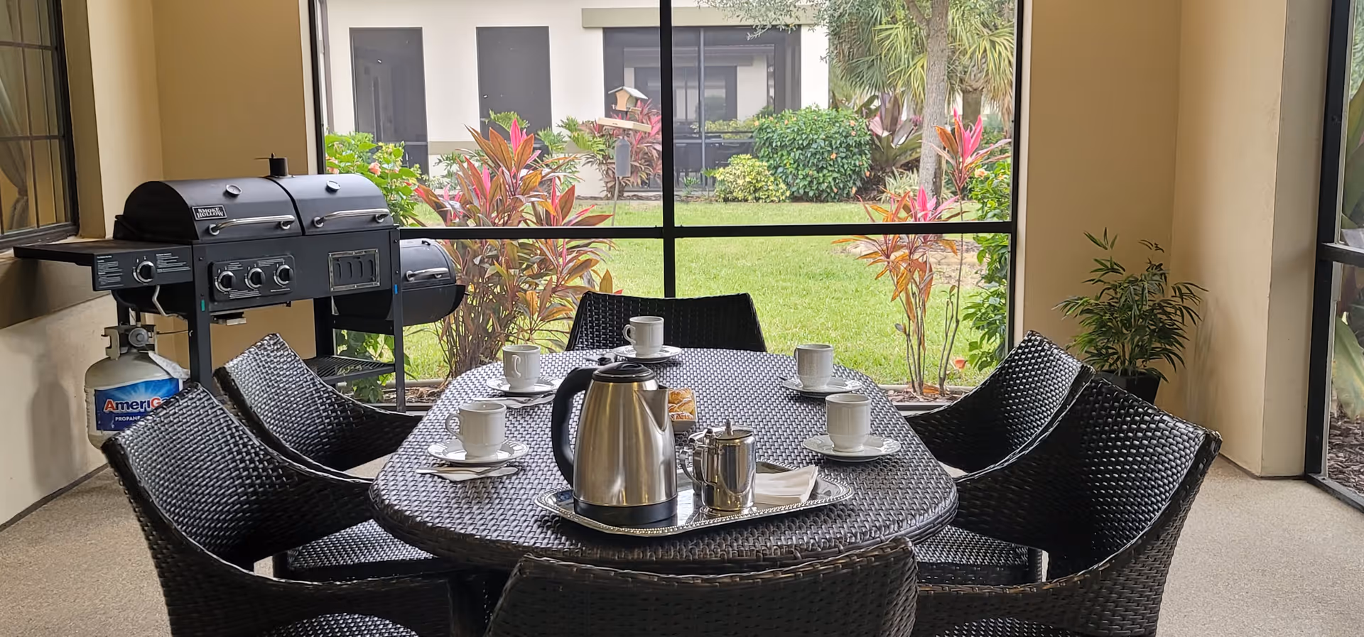 A covered outdoor patio area with a round wicker table set for coffee, including a stainless steel coffee pot, sugar container, and six white cups with saucers. Surrounding the table are six matching wicker chairs. In the background, there is a large grill and a view of a green lawn with colorful plants and bushes.