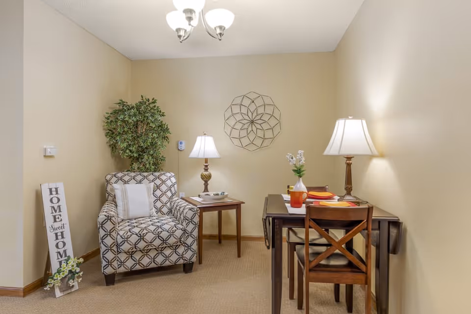 Small furnished living/dining nook with a patterned armchair, side table, small dining table and two chairs, lamps, and wall decor against beige walls.
