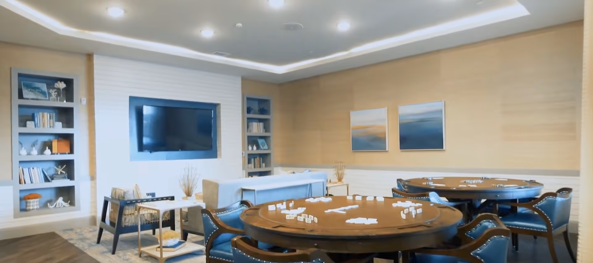 A cozy common area with two round wooden tables set up for a game with dominoes, surrounded by blue cushioned chairs. In the background, there is a light-colored sofa, built-in shelves with books and decorative items, a wall-mounted flat-screen TV, and two abstract paintings on the wall.