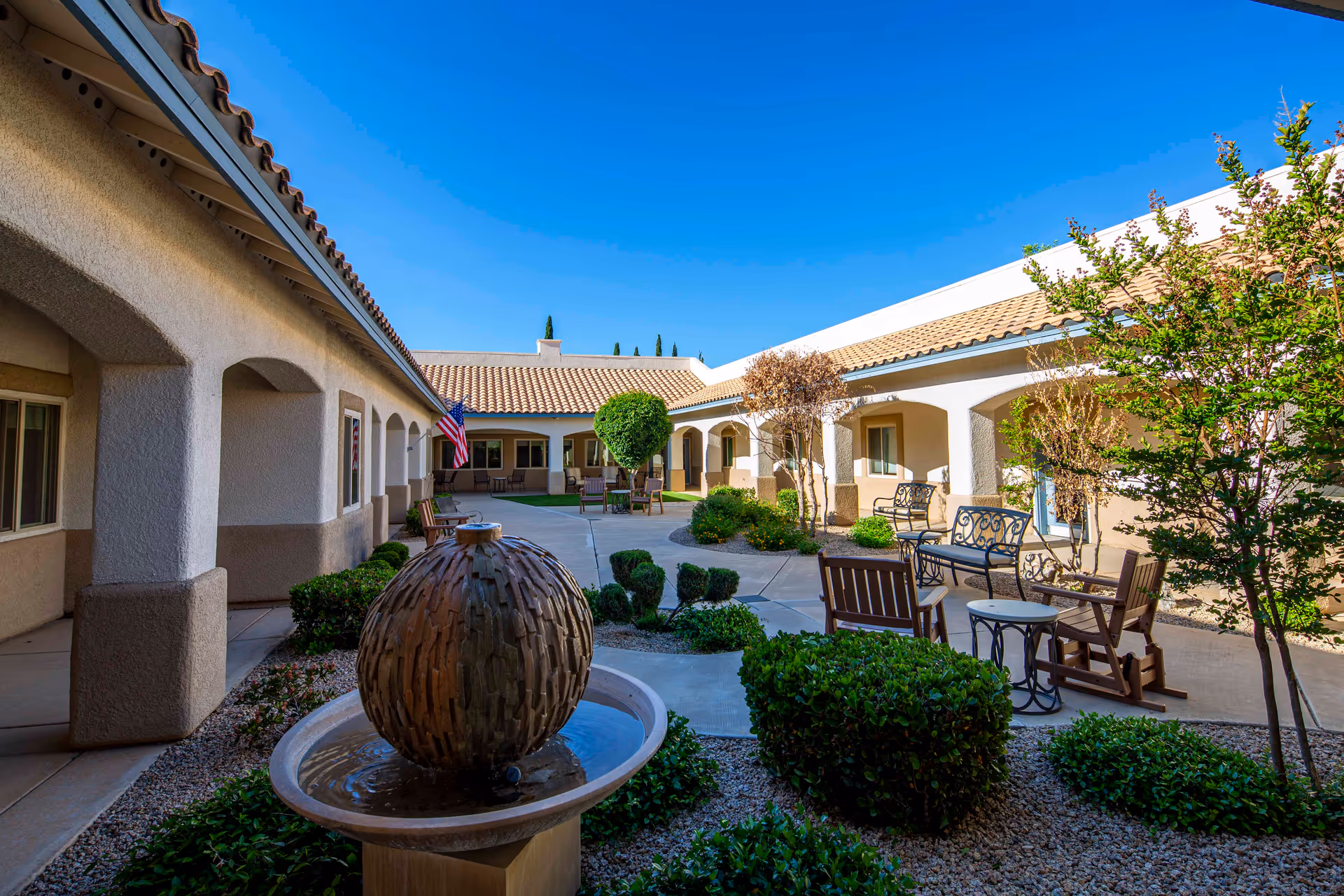 Sunny courtyard with a decorative fountain, outdoor seating, and arched walkways between single-story buildings.