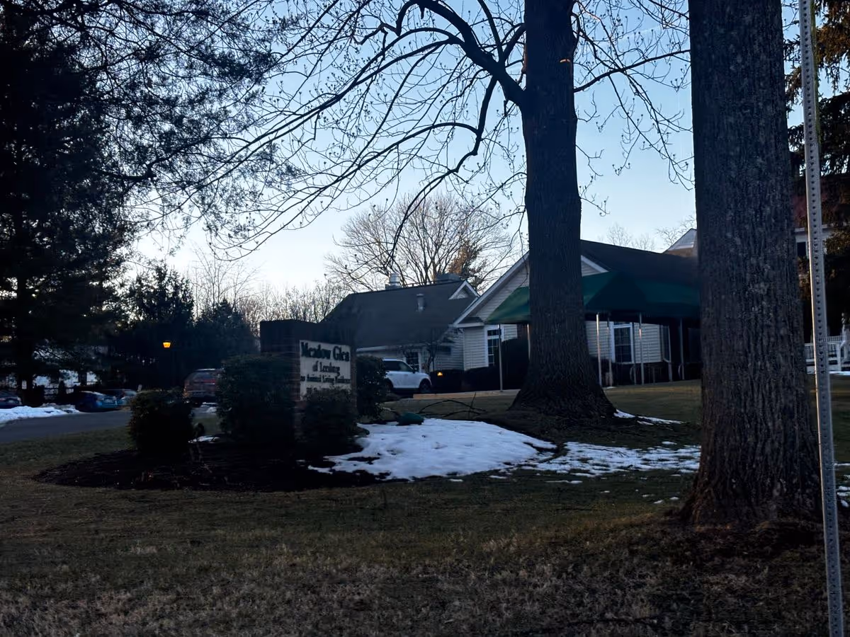 Front exterior of a senior living building with entrance awnings, a driveway, trees, a sign, and patches of snow on the lawn.