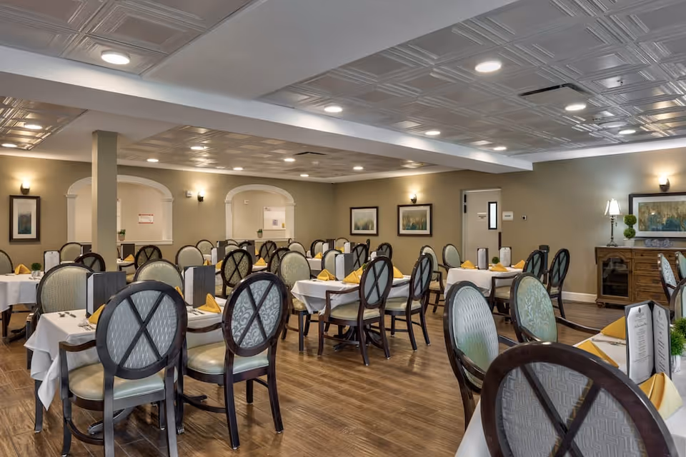 A spacious dining room with multiple tables covered in white tablecloths, each set with yellow folded napkins and menus. The room has wooden floors, beige walls with framed artwork, and a ceiling with recessed lighting and decorative panels. Chairs with cushioned seats and backs surround the tables.