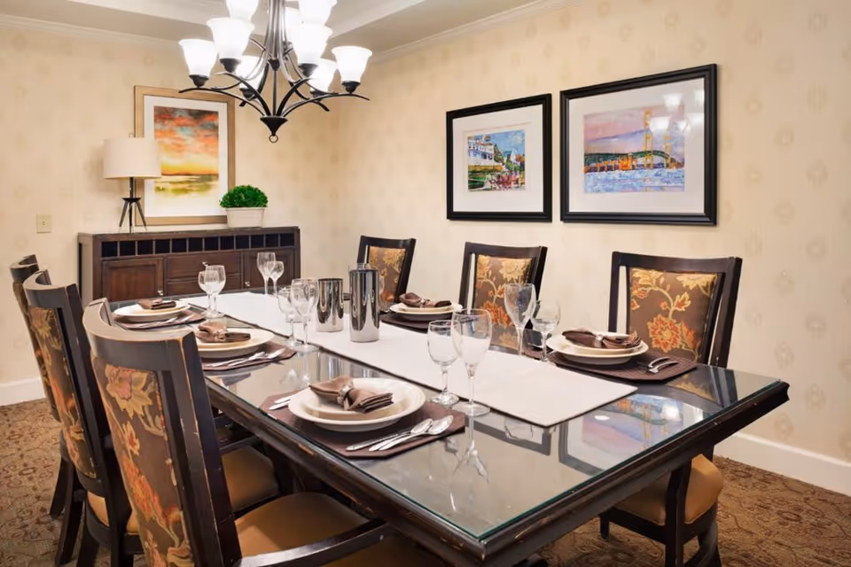 A formal dining room with a glass-top table set for six people. The table is arranged with plates, brown napkins, silverware, and multiple wine glasses. The chairs have floral upholstery. On the wall behind the table are two framed paintings, and a sideboard with a lamp, a framed picture, and a small potted plant is visible. A chandelier with multiple lights hangs above the table.