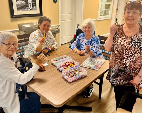 Four elderly women sitting and standing around a wooden table engaged in a bead crafting activity. The table has containers filled with colorful beads and crafting materials. The room has light-colored walls and a door in the background.