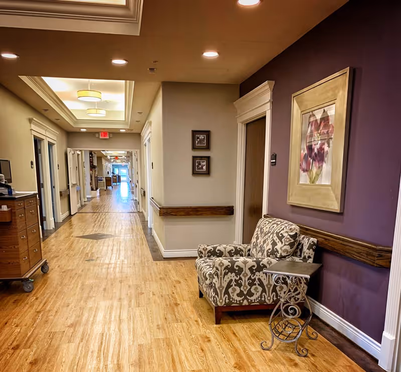 A well-lit assisted living hallway with wood floors, an upholstered chair and side table against a purple wall, framed art, and a long corridor of doors.