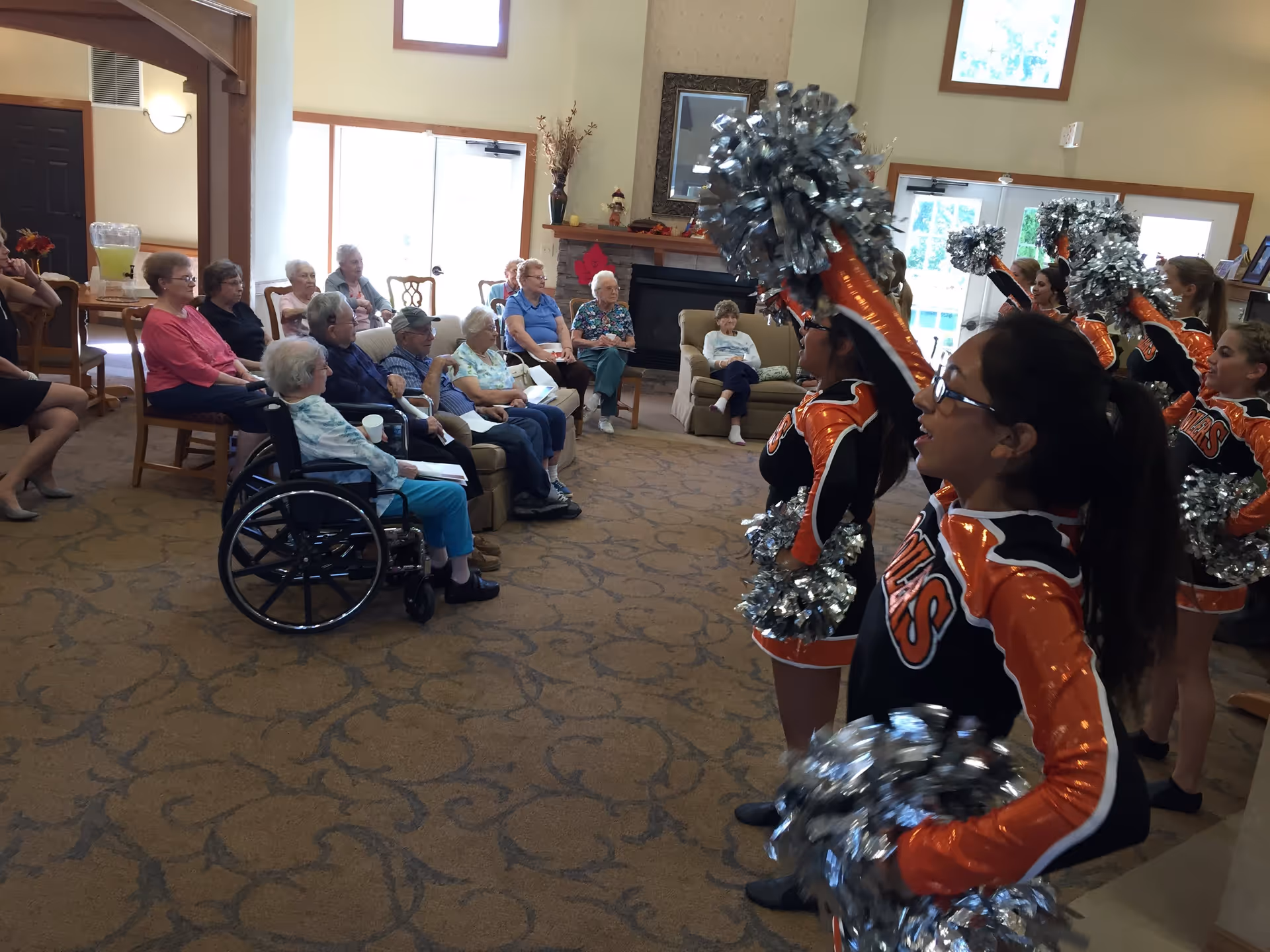 A group of elderly people seated in a common room watching a cheerleading performance by young girls in orange and black uniforms holding silver pom-poms. The room has large windows, a fireplace, and comfortable seating.