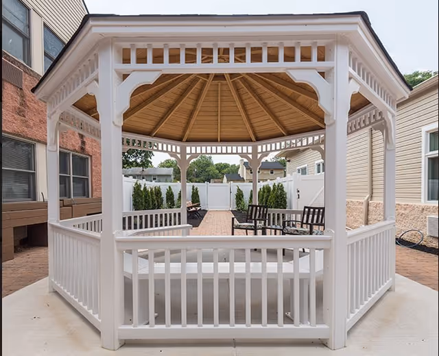 White wooden gazebo with a peaked roof in a fenced courtyard furnished with chairs and potted shrubs.