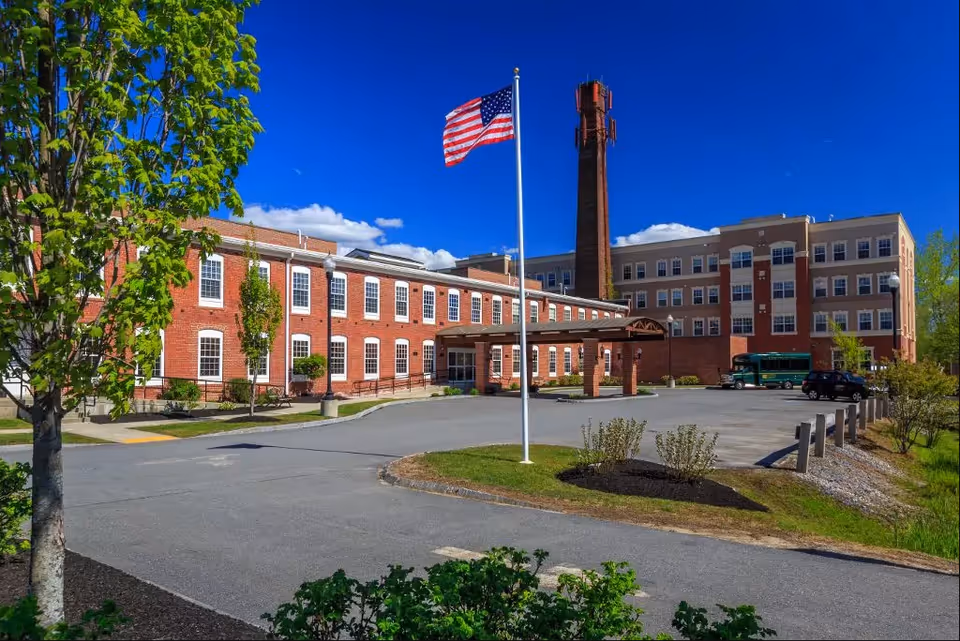 Exterior view of the American House Keene facility showing a large brick building with multiple windows, a covered entrance, an American flag on a flagpole, a tall brick smokestack, a parking area with vehicles, and surrounding greenery under a clear blue sky.