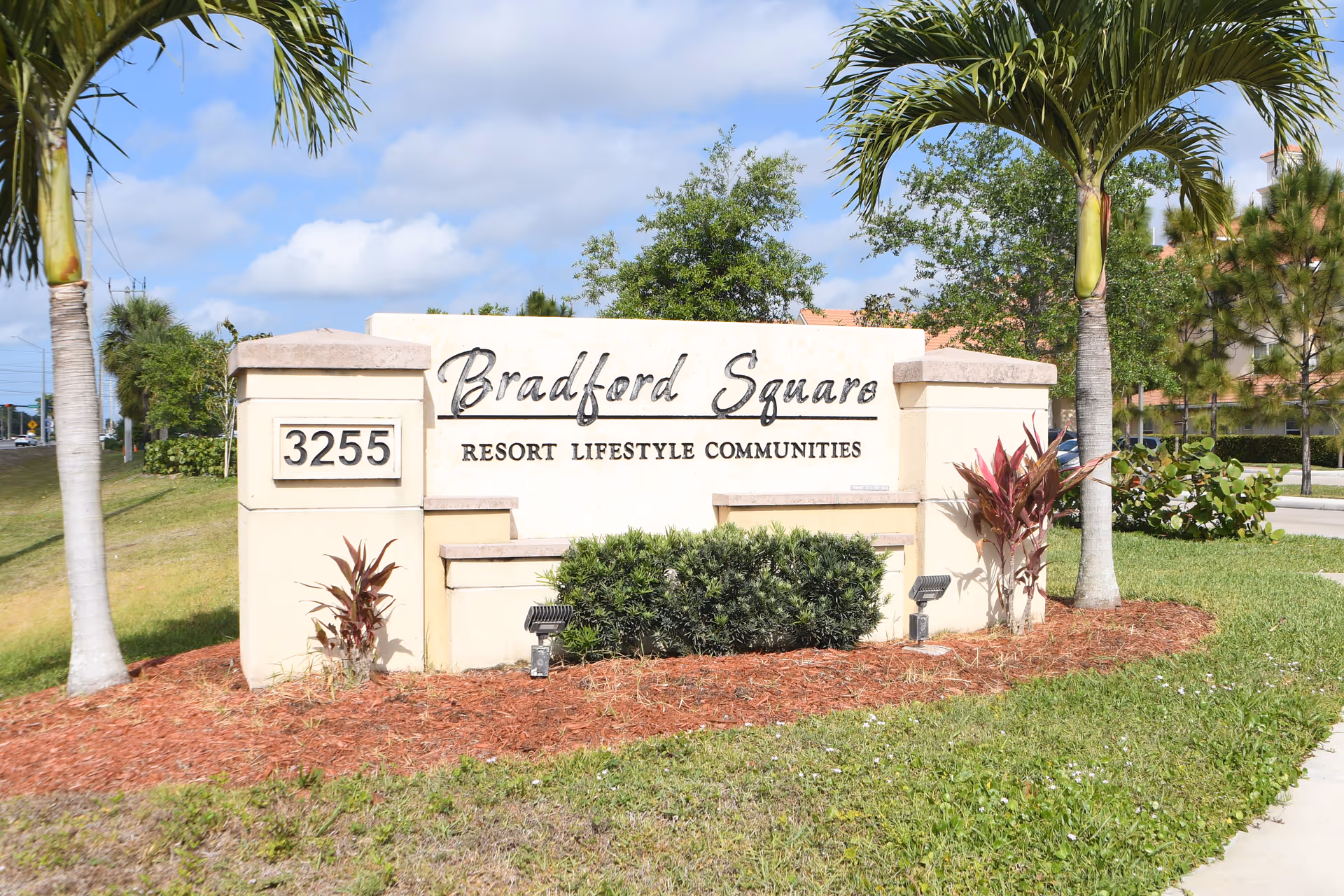 Outdoor view of a large stone sign for Bradford Square Resort Lifestyle Communities with the address number 3255, surrounded by palm trees, bushes, and grass under a partly cloudy sky.