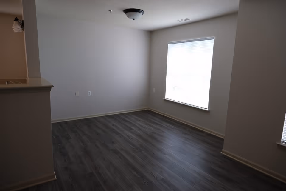 Empty interior living area with dark wood-look flooring, a large window with blinds, neutral walls, and a partial kitchen counter visible on the left.