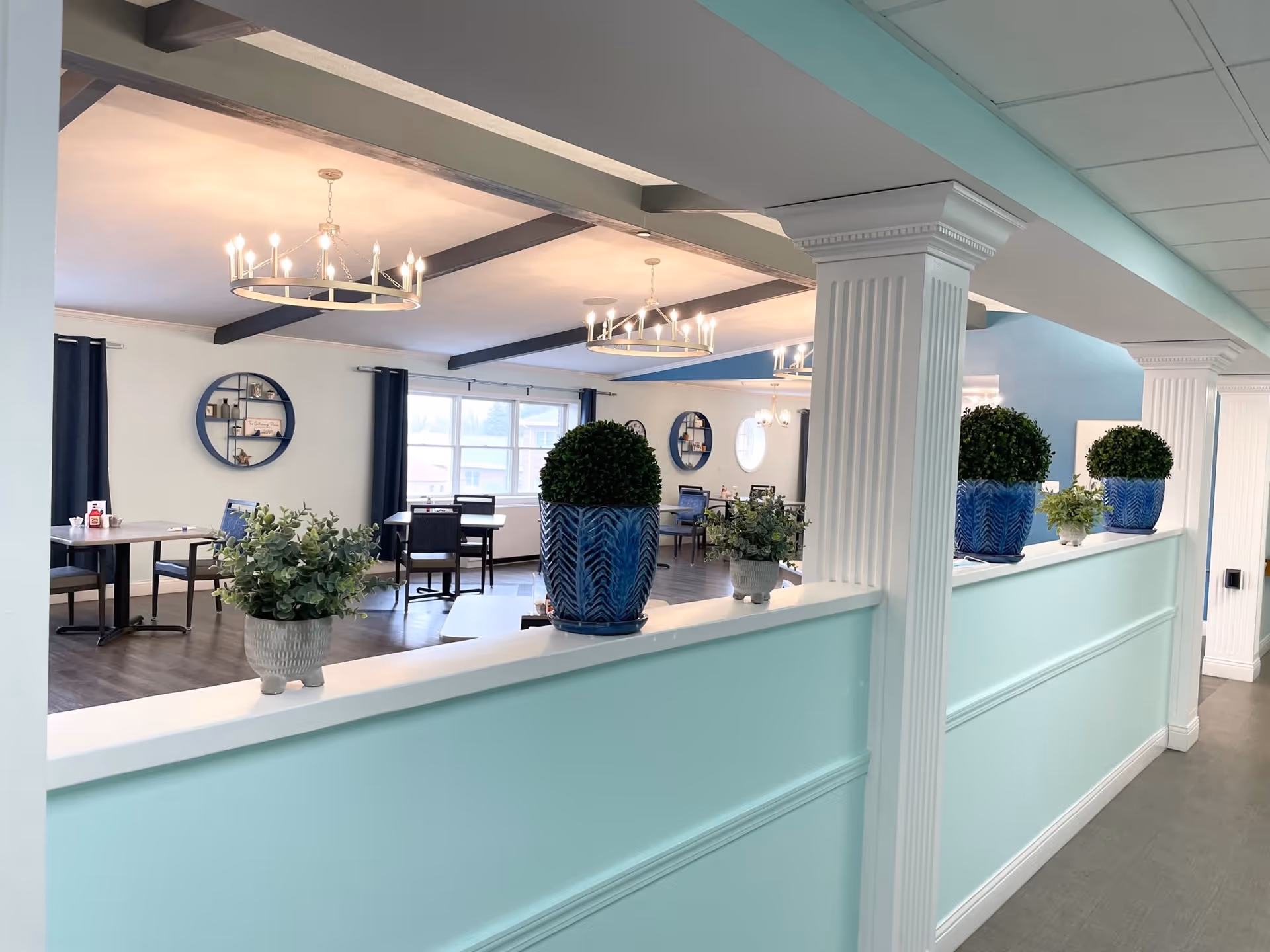 Bright communal dining/activity room with tables, chandeliers, columns, and potted plants on a teal half-wall.