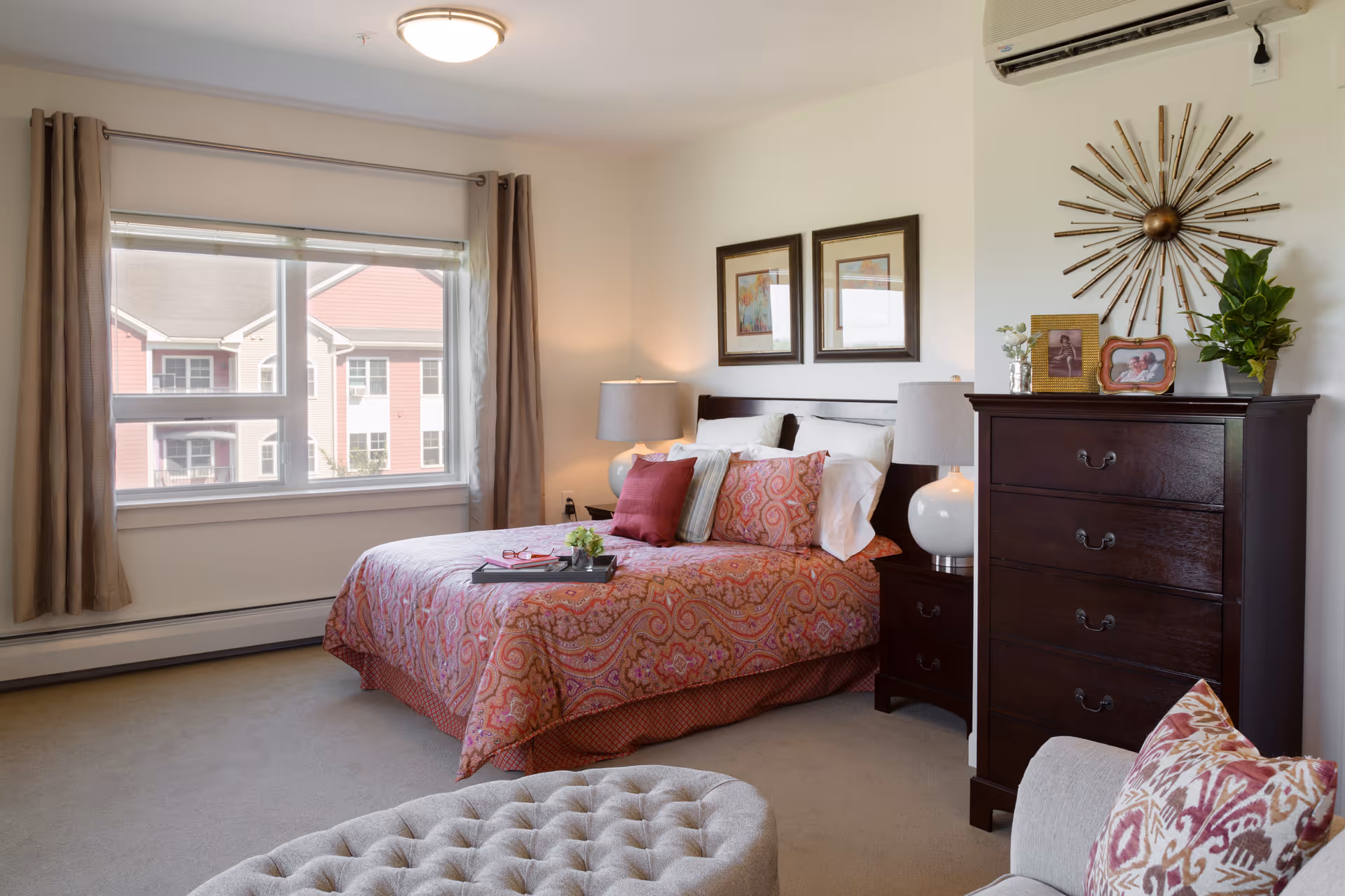 A well-lit bedroom with a large window showing an exterior view of a building. The room features a bed with a colorful patterned bedspread and multiple pillows, two bedside tables with lamps, a dark wooden dresser adorned with framed photos, a plant, and a decorative sunburst wall art above it. There is also a beige upholstered ottoman and a chair with a patterned cushion in the foreground.