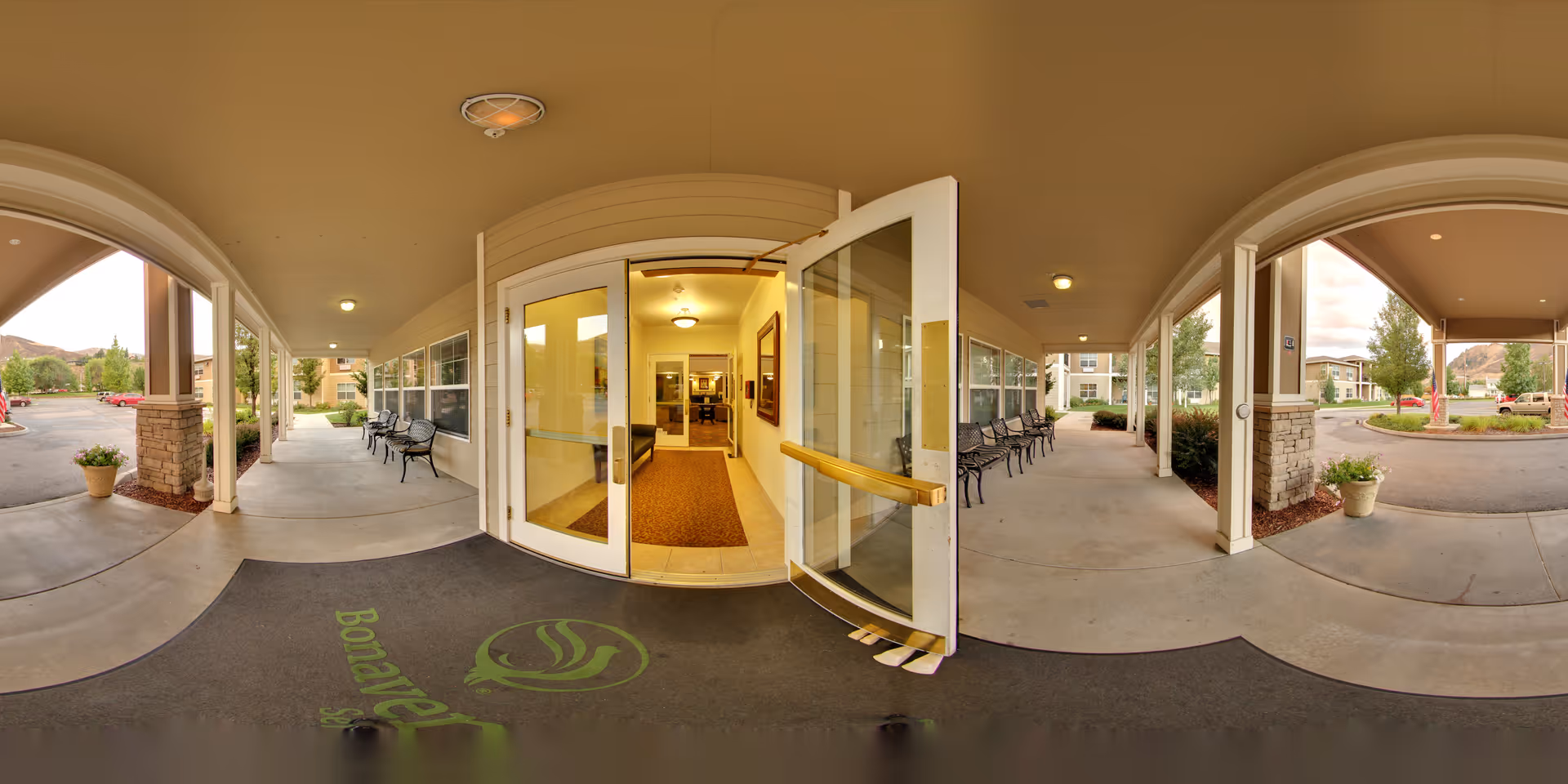 Covered entrance area of Wenatchee Senior Living by Cogir with glass double doors, benches along the walls, potted plants, and a view of the parking lot and surrounding buildings.