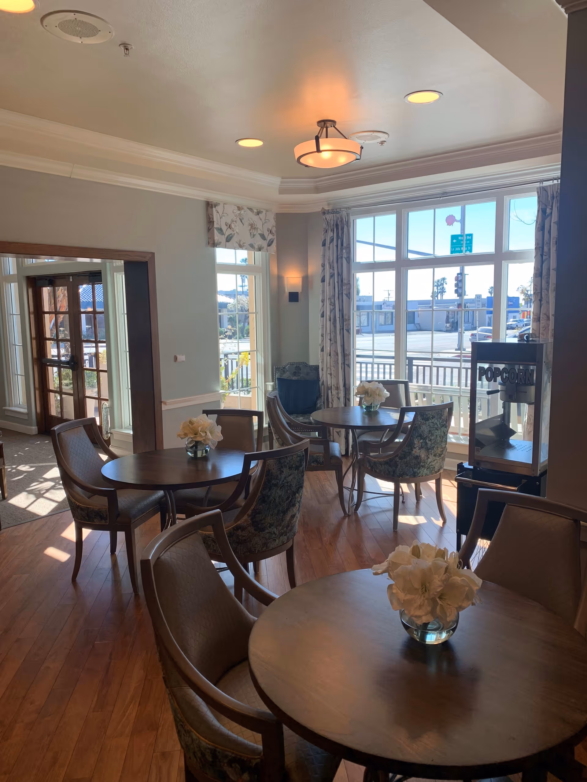 A bright and cozy common area with round wooden tables and cushioned chairs arranged around them. Each table has a small vase with white flowers. Large windows let in natural light and offer a view of the street outside. There is a popcorn machine near the window and floral curtains and valances adorn the windows. The room has wooden flooring and soft ceiling lighting.