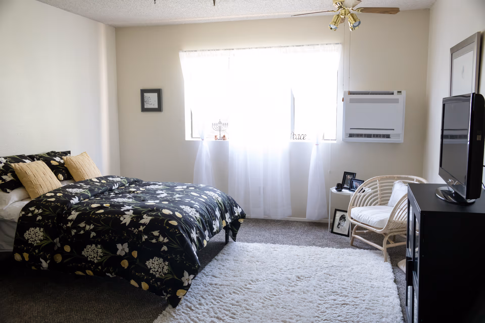 A bright bedroom with a bed covered in a black floral comforter and two beige pillows. There is a white fluffy rug on the floor, a wicker chair with a white cushion, a small side table with framed photos, and a flat-screen TV on a black stand. A window with sheer white curtains lets in natural light, and a wall-mounted air conditioning unit is visible.
