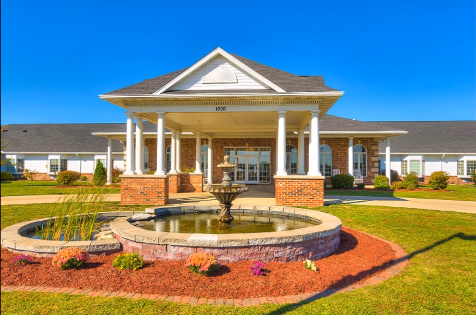 Front exterior view of Cedarhurst Senior Living of Godfrey featuring a covered entrance with white columns, a brick facade, a circular fountain in the foreground, and well-maintained landscaping with flowers and grass under a clear blue sky.