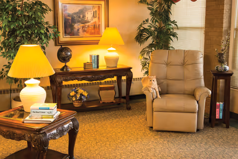 A cozy living room area with a beige recliner chair where a light orange cat is sitting. The room features two wooden side tables with lamps, a wooden console table with books, a globe, and a flower arrangement. There are large green plants and a framed painting on the wall, with window blinds partially closed.