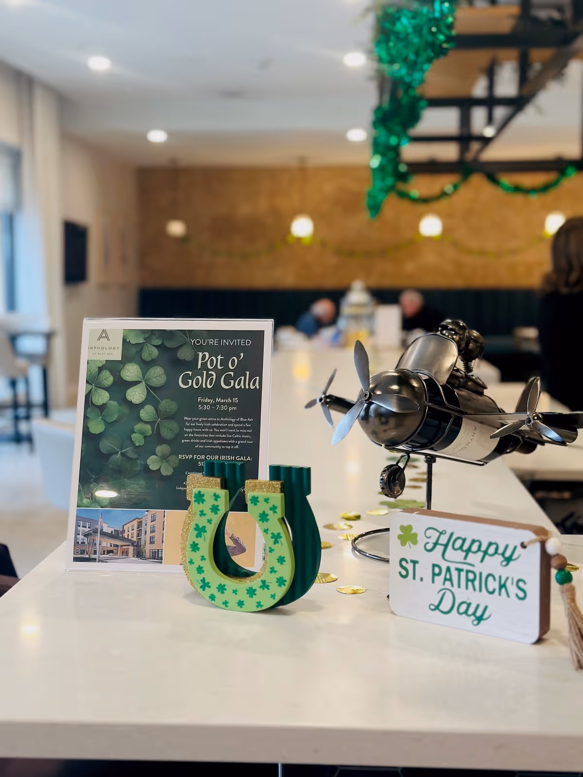 Countertop display with St. Patrick's Day decorations, a "Pot o' Gold Gala" invitation, a decorative horseshoe and a small metal airplane in a communal dining area.