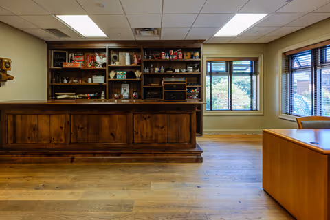 Interior common area featuring a large wooden bar/counter with shelving stocked with items, hardwood floors, and windows.