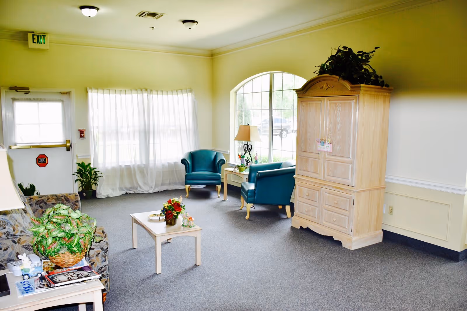 A bright and cozy living room area with a large window letting in natural light. The room features two teal armchairs, a beige wooden cabinet with a plant on top, a small coffee table with a flower arrangement, and a patterned sofa with a large green plant on the side table. The walls are painted light yellow with white trim, and there is an exit door with a window and a sheer curtain-covered window beside it.