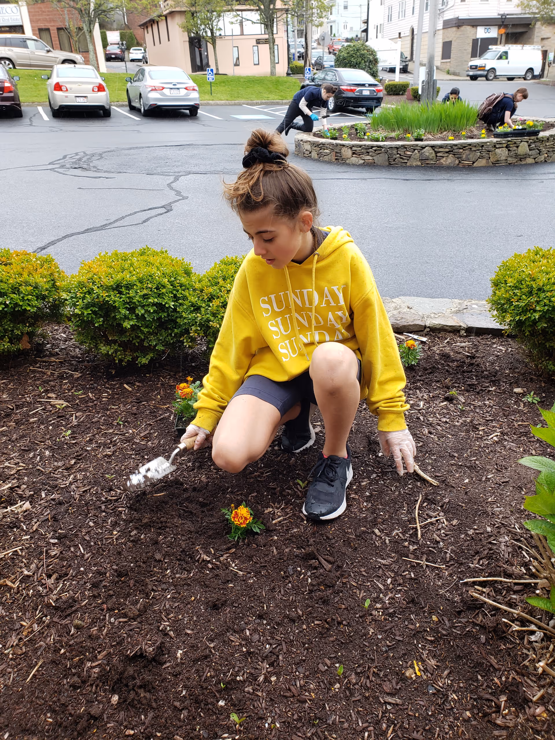 A young girl wearing a yellow hoodie with the word 'SUNDAY' printed on it is kneeling on the ground, planting flowers in a garden bed. She is holding a small gardening tool and wearing gloves. In the background, two other people are also gardening near a stone-bordered flower bed in a parking lot area with several parked cars and buildings.