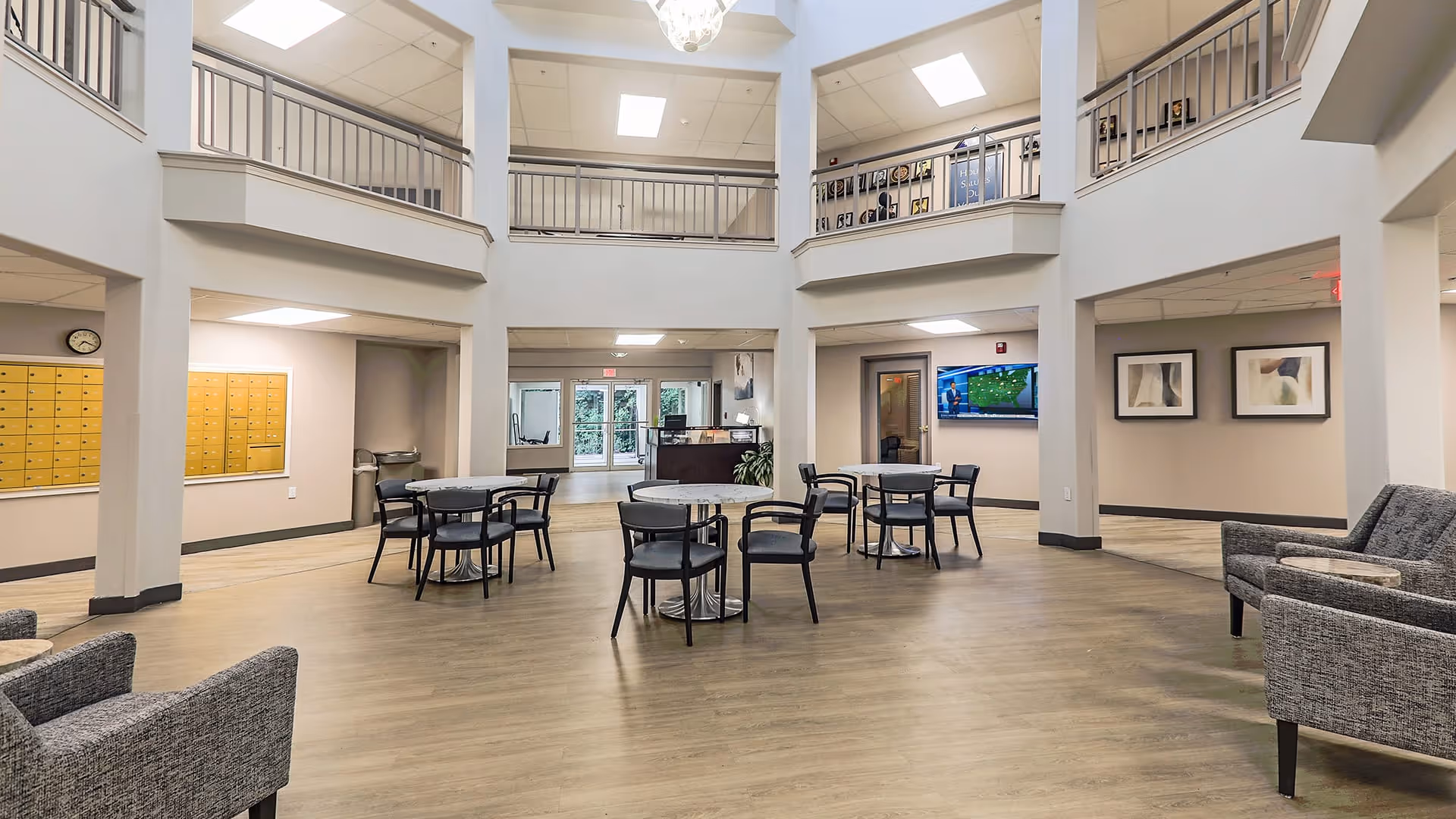 Spacious and well-lit common area in a senior living facility with high ceilings and a second-floor balcony. The room features several round tables with chairs, gray upholstered armchairs, a wall-mounted TV, framed artwork, and a set of mailboxes on one wall. The flooring is wood-style, and there are large windows and glass doors allowing natural light to enter.