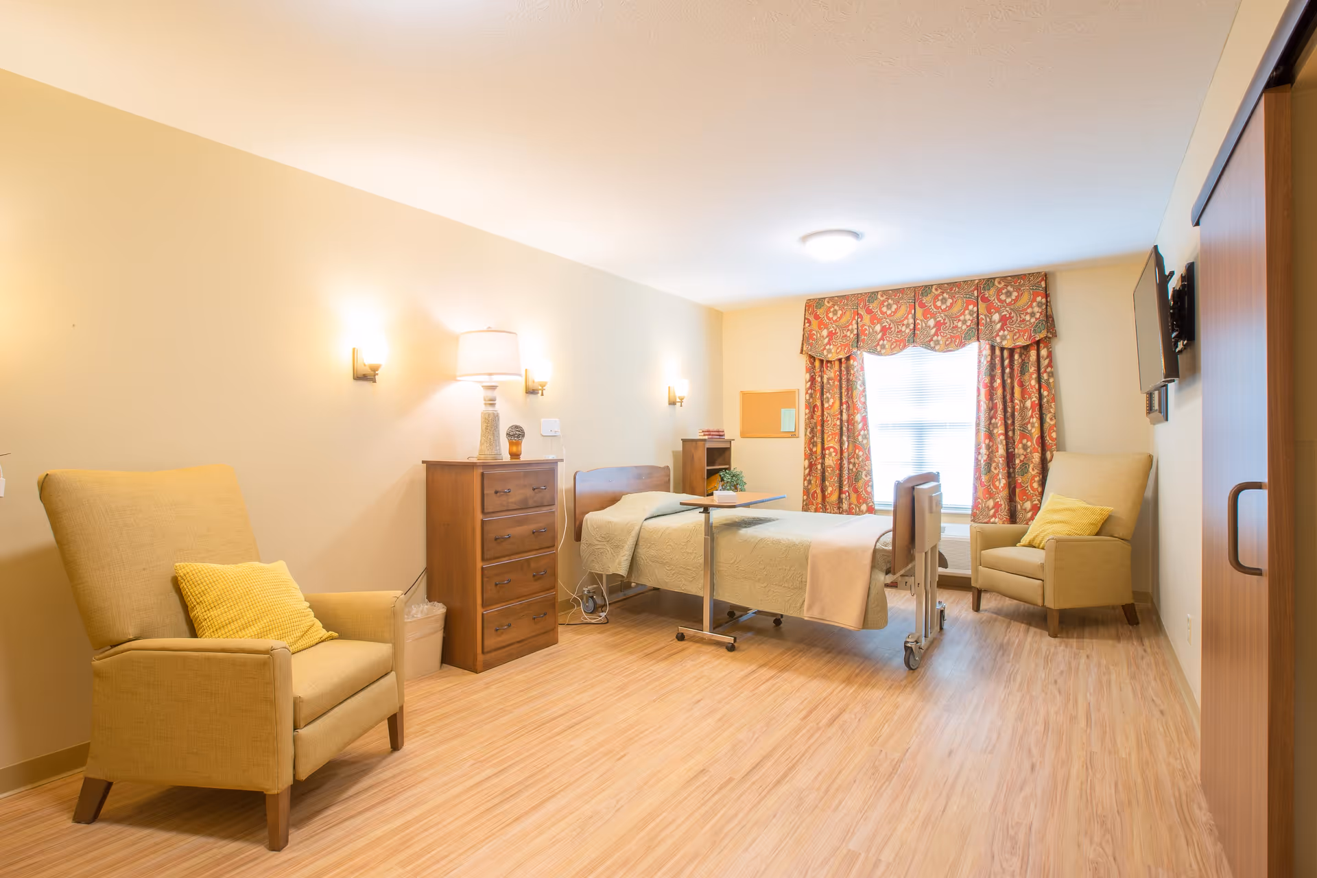 A well-lit senior living bedroom with a hospital-style bed, two beige armchairs with yellow pillows, a wooden dresser with a lamp, a small table on wheels, and a window with floral curtains. The room has light wood flooring and neutral-colored walls.