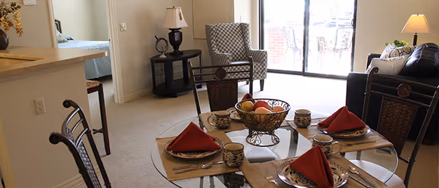 Interior view of a senior living facility showing a dining area with a glass table set for four with plates, cups, and red napkins. In the background, there is a living room with a black leather sofa, a patterned armchair, a side table with a lamp, and large sliding glass doors leading to an outdoor patio.