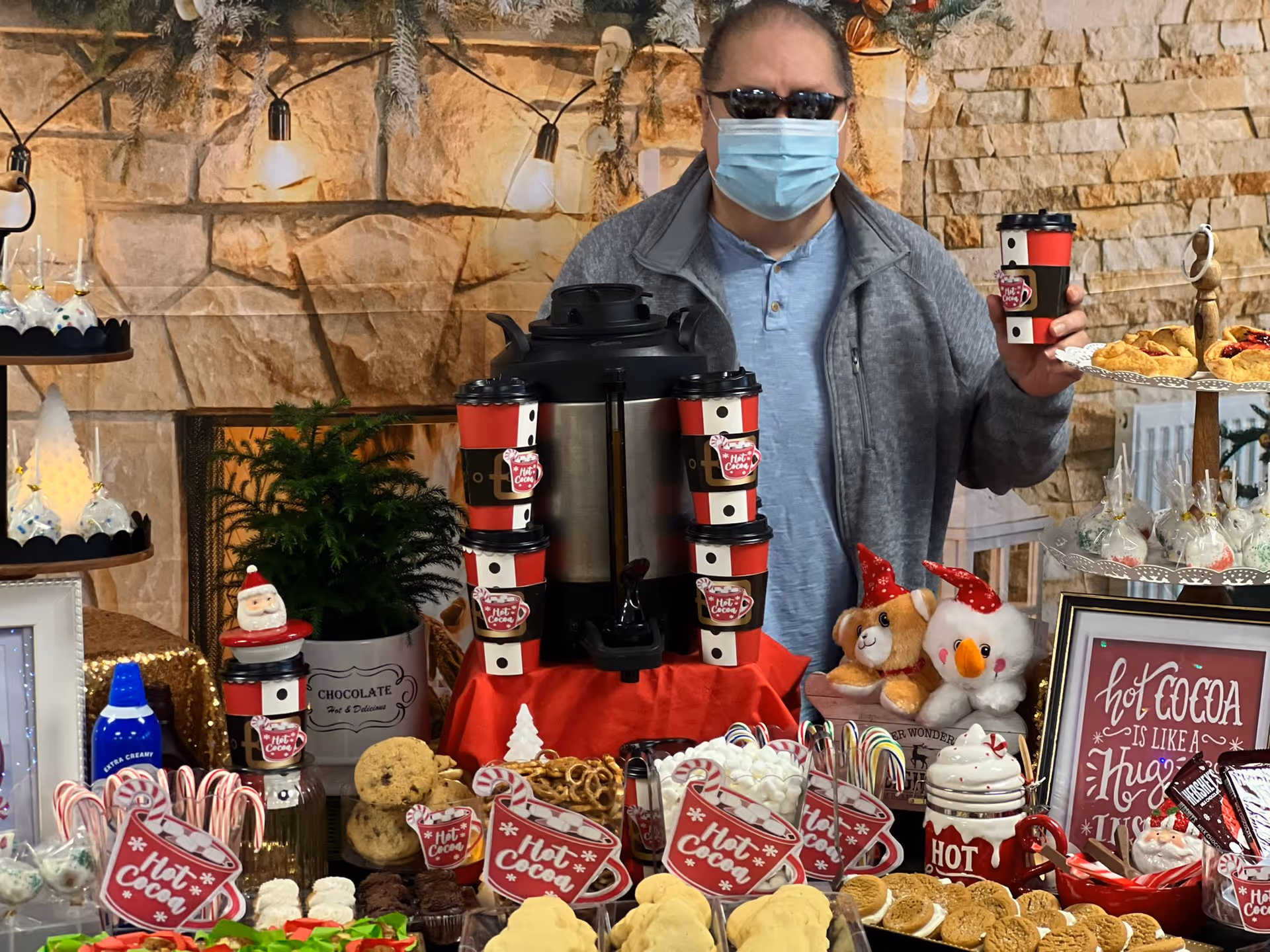 A person wearing sunglasses and a face mask stands behind a festive hot cocoa and dessert table decorated with holiday-themed items including candy canes, cookies, marshmallows, and stuffed animals wearing red hats. The background features a stone wall with string lights and greenery.