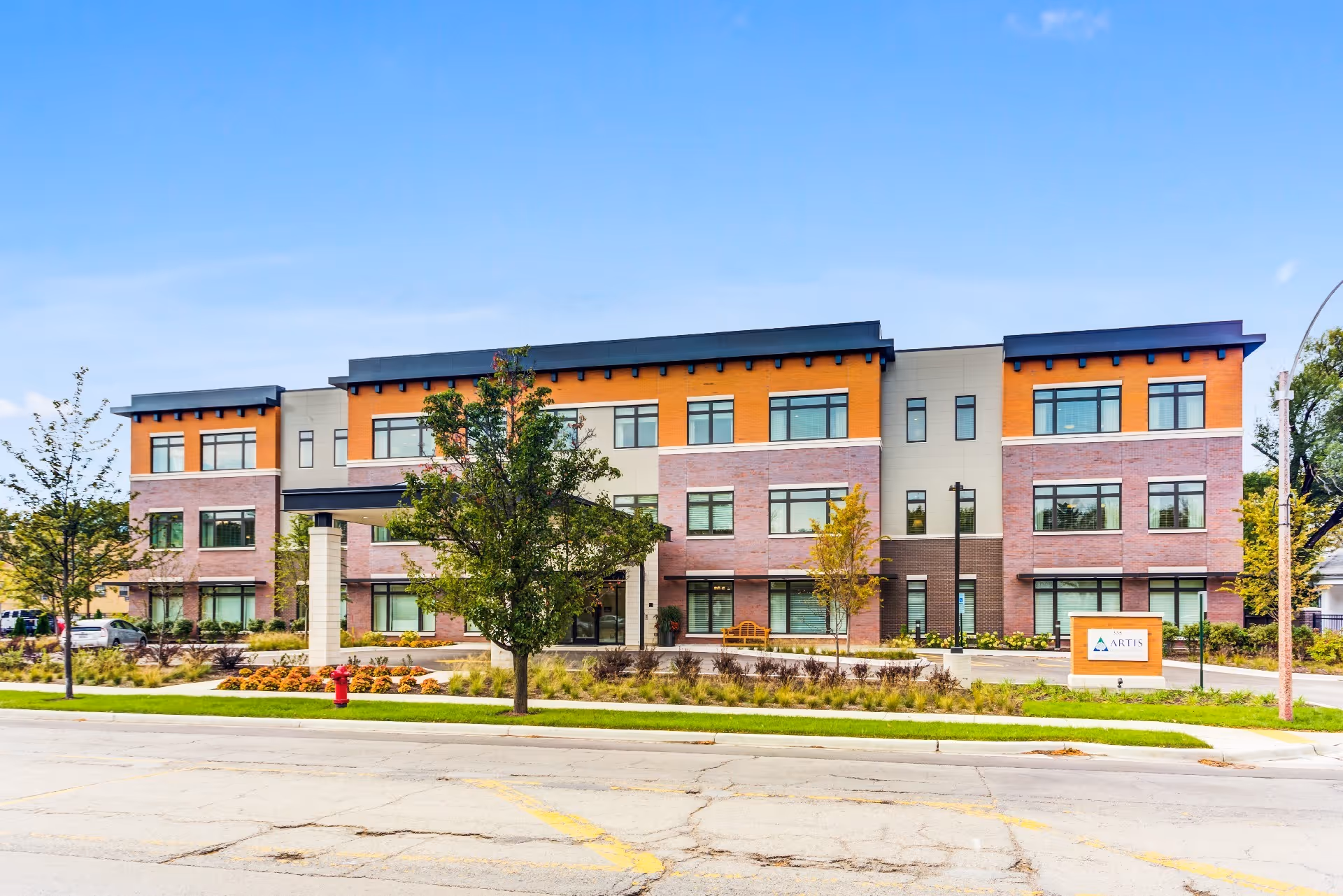 Front exterior view of a modern three-story senior living facility building with a mix of orange, beige, and brick facade, several windows, trees, and landscaping in front, under a clear blue sky.