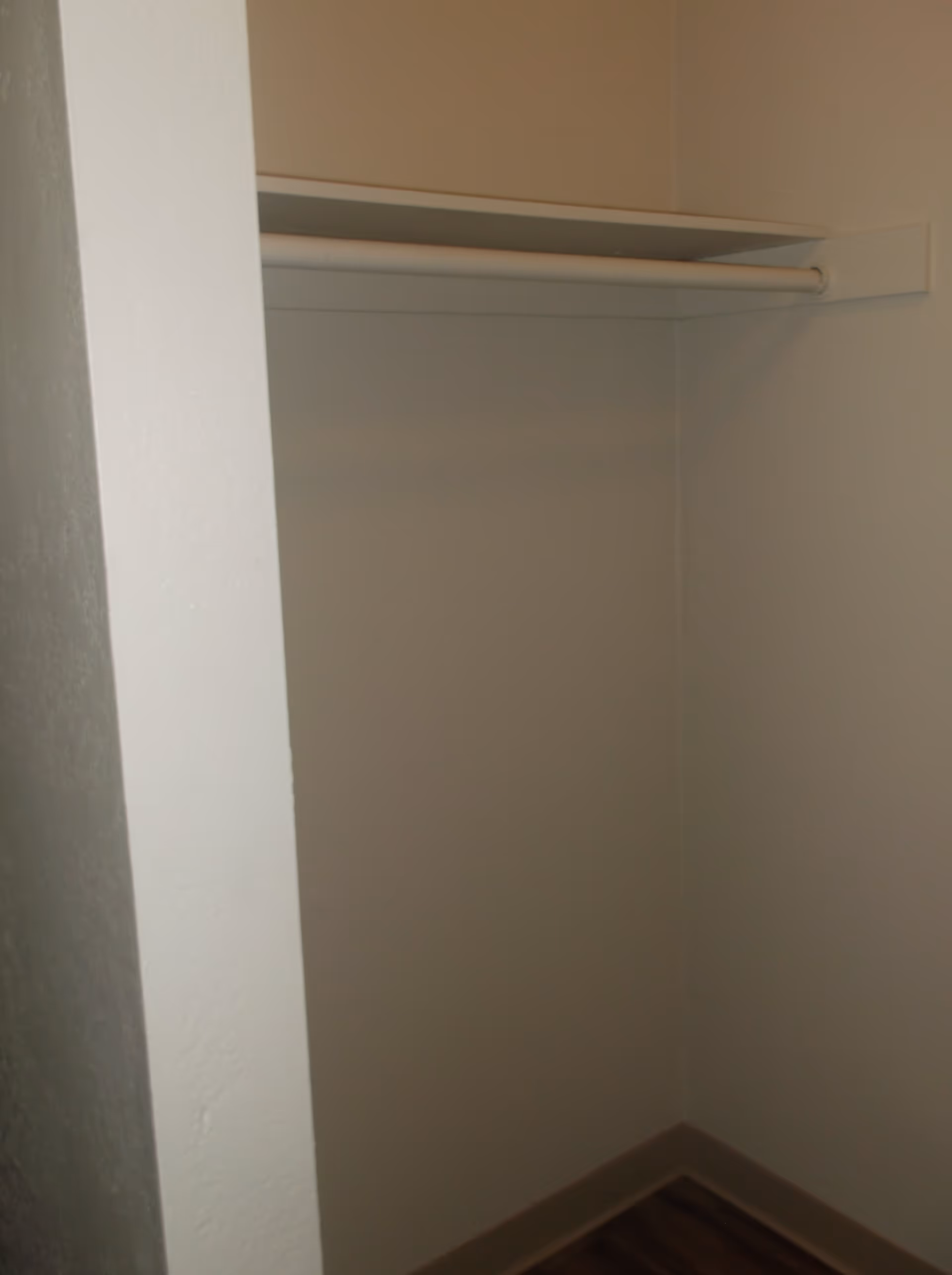 Empty closet with a single white hanging rod and a shelf above it, surrounded by plain white walls and a wooden floor.