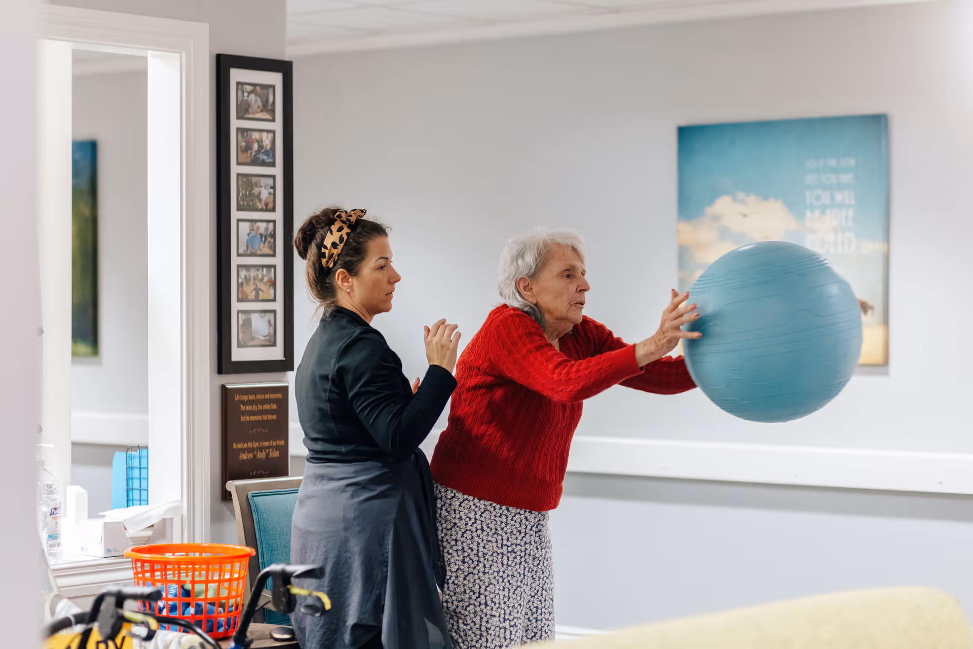 An elderly woman in a red sweater and floral skirt is holding a large blue exercise ball while a younger woman stands behind her, assisting with the activity in a room with light gray walls and framed pictures.