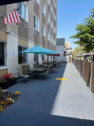 Outdoor patio area alongside a multi-story brick building with several tables and chairs under blue and red umbrellas. An American flag is mounted on the building wall, and there are flower beds with colorful flowers near the seating area. A fence runs along the right side of the patio, with hanging flower baskets attached to it. The sky is clear and blue.
