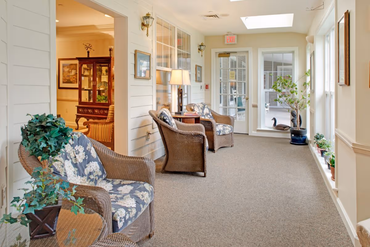 A bright and cozy indoor sitting area with three wicker chairs featuring floral cushions arranged along a wall with windows. A small table with a lamp and books is placed between two chairs. The room has beige carpeting, white walls, and several potted plants near large windows that let in natural light. A glass door leads to an outdoor patio area with more plants and decorative items visible outside.