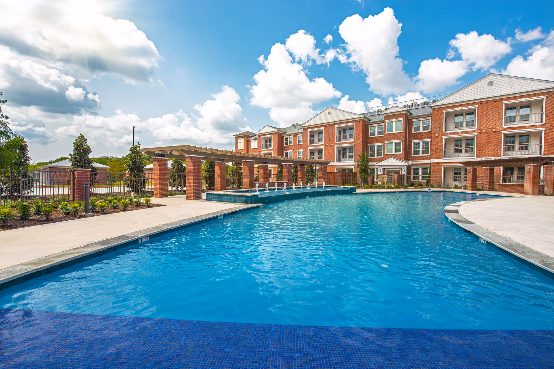 Outdoor swimming pool with clear blue water in front of a three-story brick building under a bright sky with scattered clouds. The pool area features a pergola with brick columns and small water fountains along the edge of the pool.