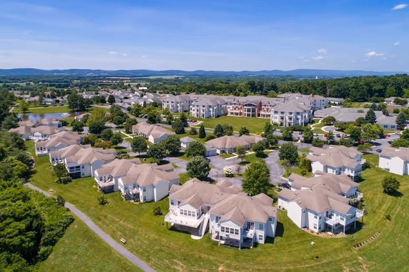 Aerial view of Buckingham's Choice senior living community showing multiple residential buildings with beige roofs surrounded by green lawns, trees, and walking paths under a blue sky with scattered clouds.