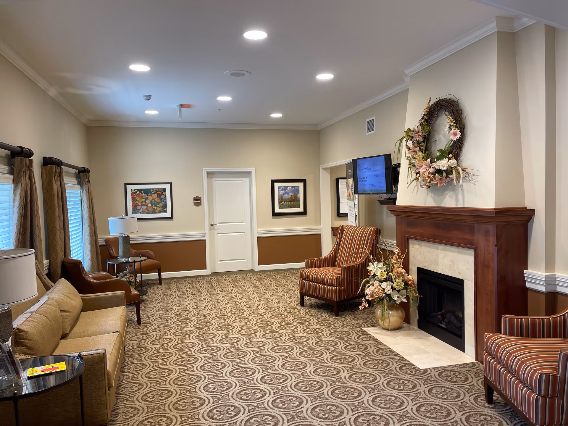 A cozy living room area in a senior living facility with beige walls and patterned carpet. The room features a beige sofa, two brown leather chairs, and two striped armchairs arranged around a wooden fireplace decorated with a floral wreath and a vase of flowers. There are framed paintings on the walls, a flat-screen TV mounted near the fireplace, and windows with curtains letting in natural light.
