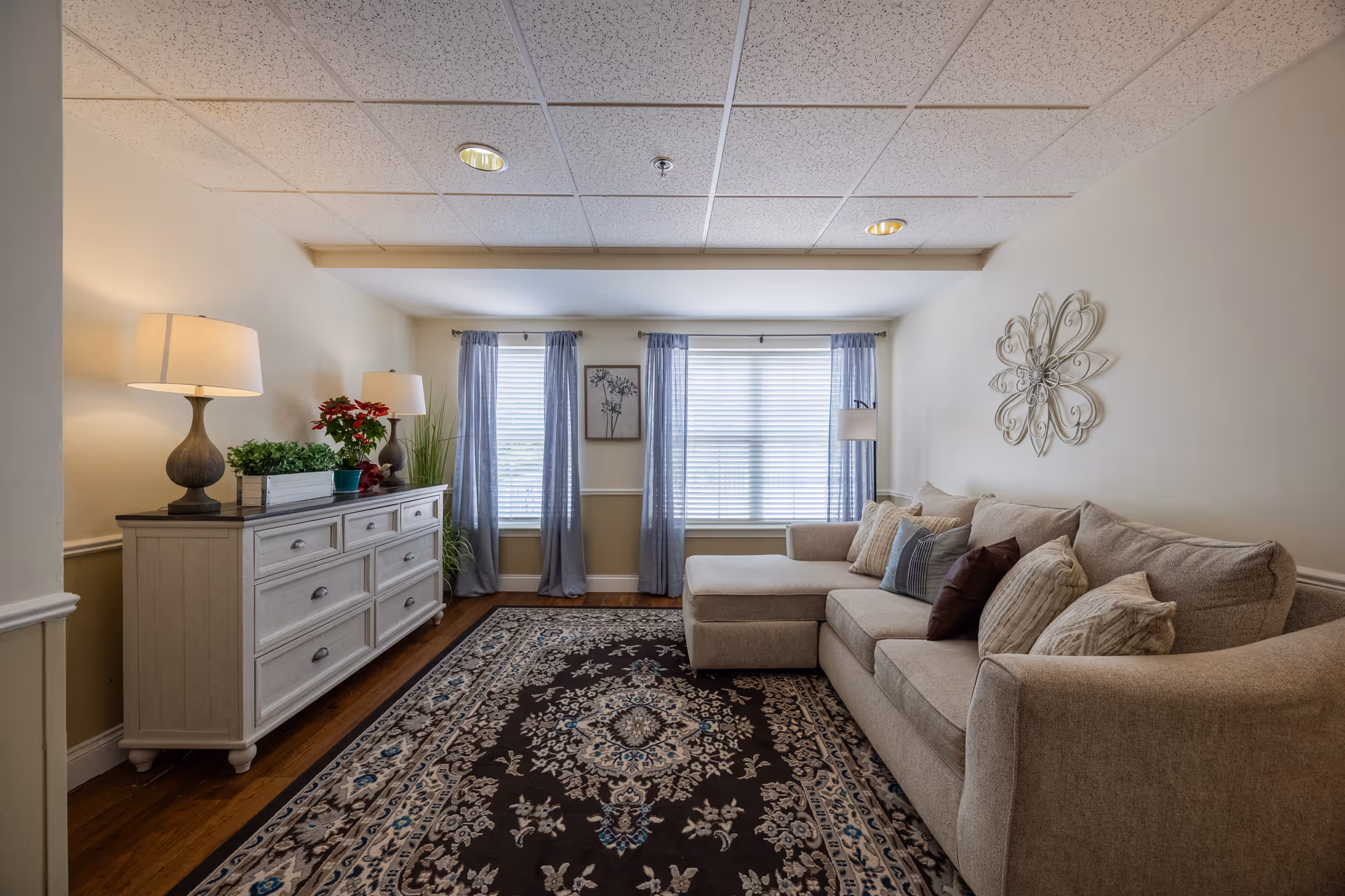 A cozy living room with a beige sectional sofa adorned with multiple pillows, a large patterned area rug, a white dresser with two table lamps and plants on top, two windows with blue curtains, and a decorative metal wall art piece.