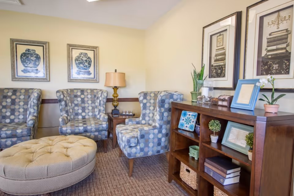 A cozy sitting area with three patterned armchairs arranged around a large beige tufted ottoman. There is a wooden side table with a lamp between two chairs. On the right side, a wooden bookshelf holds decorative items including framed pictures, small plants, and books. The walls are light yellow and decorated with framed artwork.