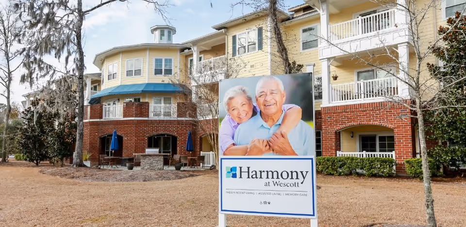 Exterior view of a senior living facility building with a large sign in the foreground showing an elderly couple smiling. The building has a combination of yellow siding and red brick with balconies and outdoor seating areas.
